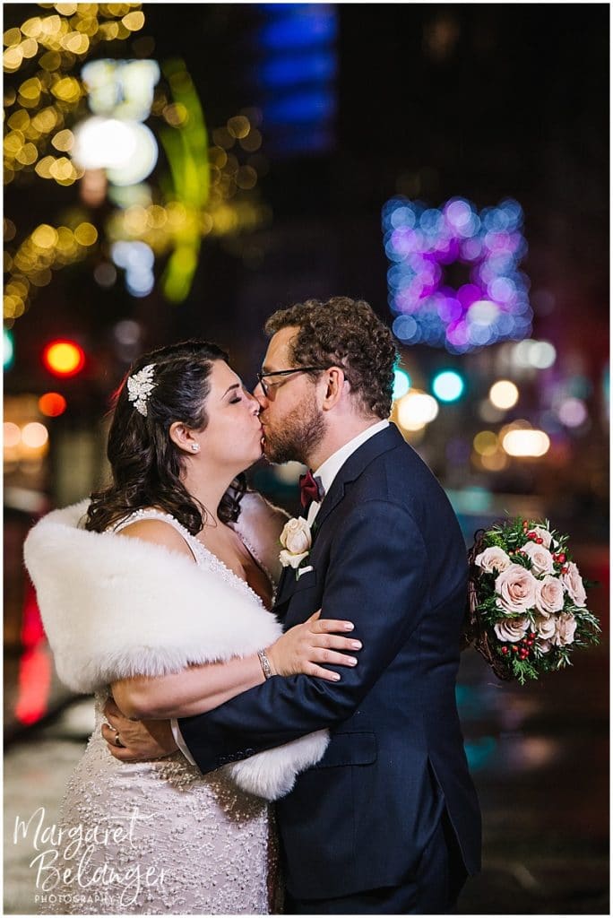 Outdoor winter wedding portraits of the bride and groom. The bride carries her bouquet and is wearing a white fur stole as they pose in front of city Christmas lights.