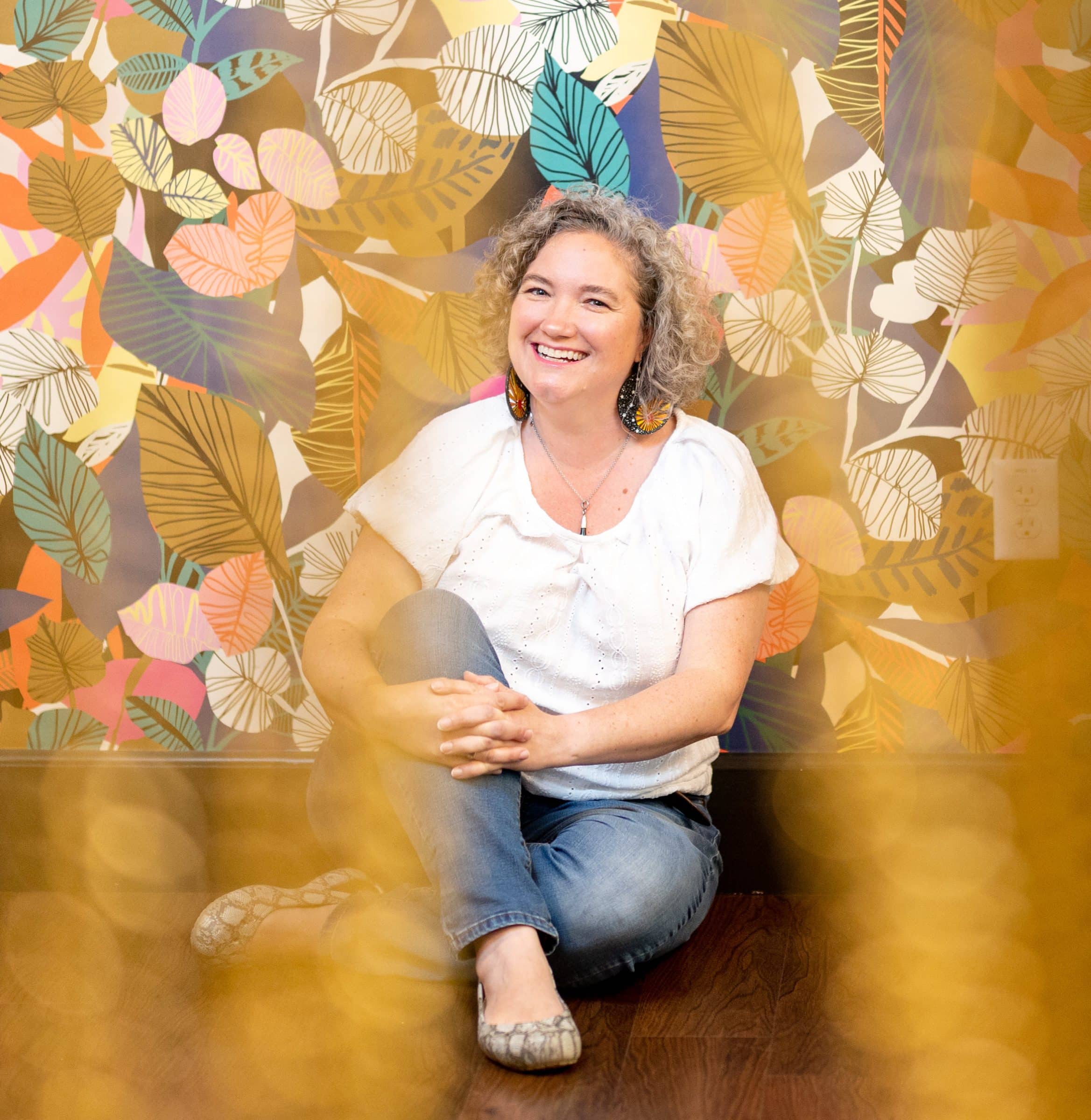 Meg Belanger sits against floral wallpaper, shot through a gold beaded curtain.