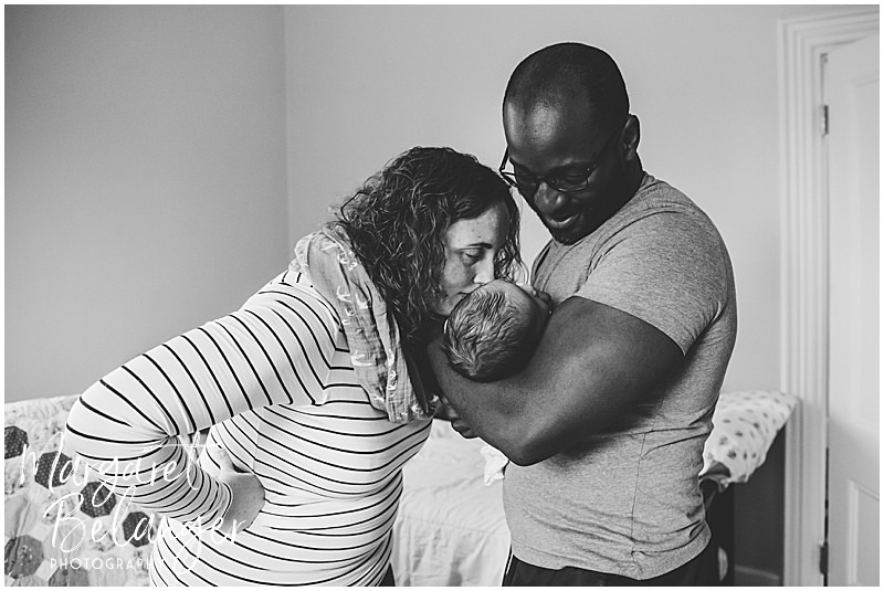 Black & white photo of dad holding newborn baby boy while mom kisses his head