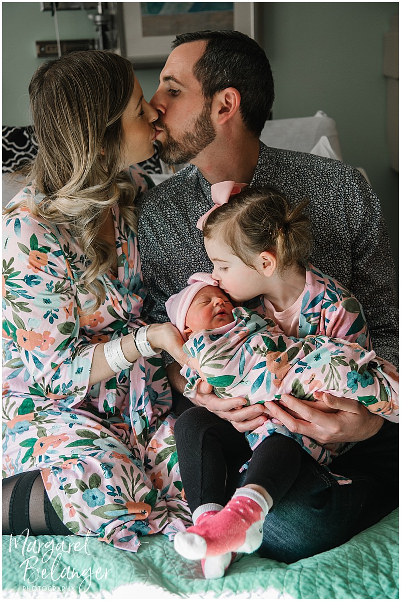 Family photo with parents kissing and big sister kissing the newborn baby