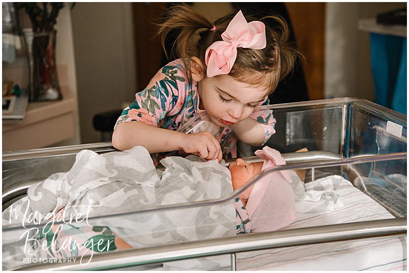 Toddler big sister peering into the bassinet to see her newborn baby sister