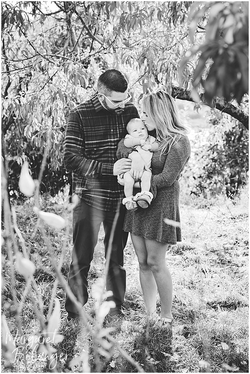B&W photo of parents holding baby, mom kissing baby's head, in an orchard at Boston Hill Farm, Andover