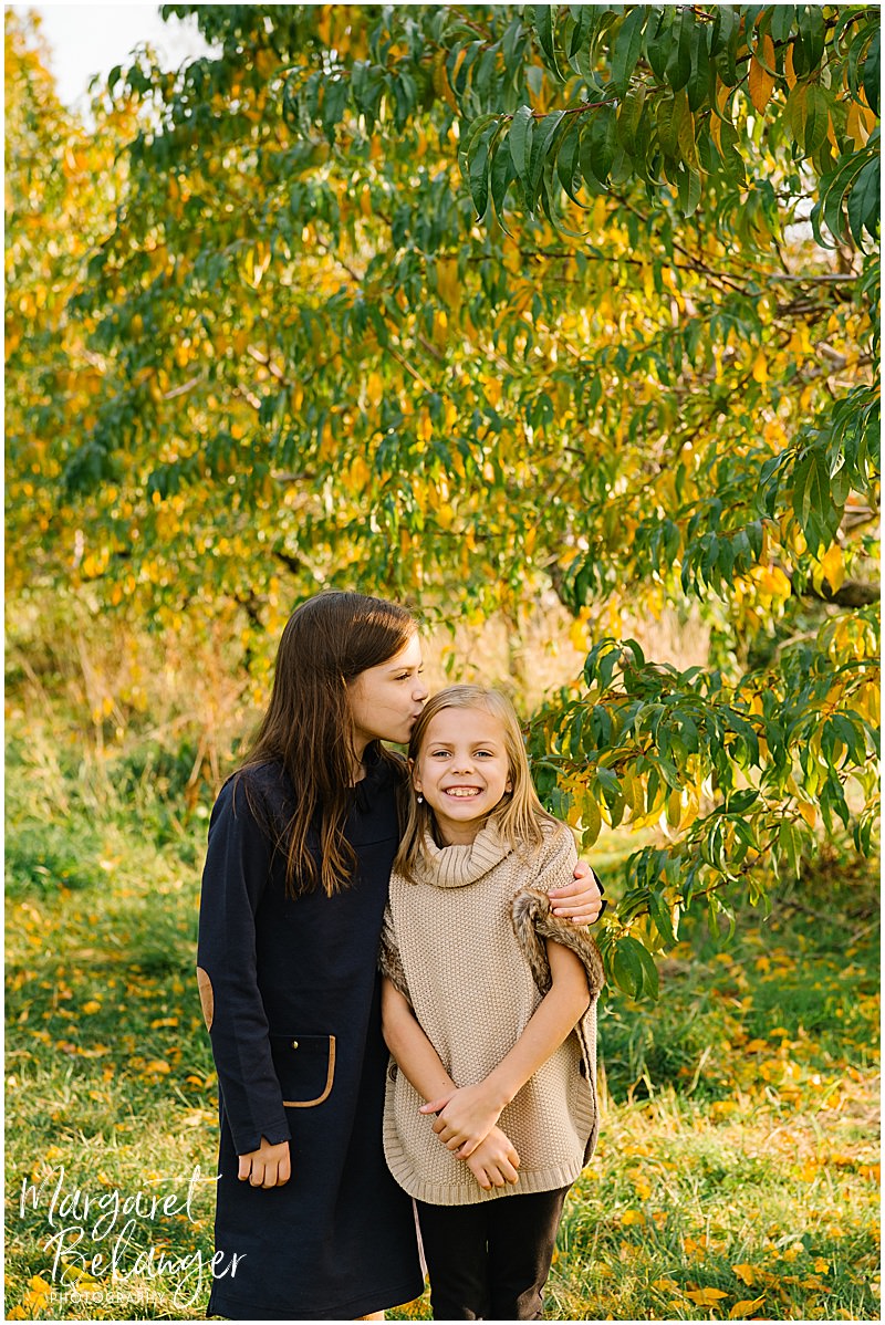 Older sister stands with younger sister and kisses her on the head in the orchard at Boston Hill Farm, Andover