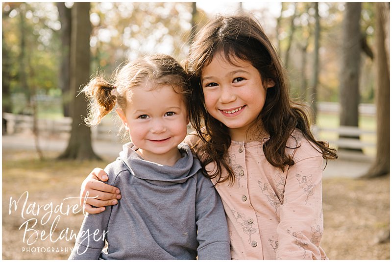Big sister puts her arm around little sister and they both smile for the camera during their fall family session at Endicott Park in Danvers.
