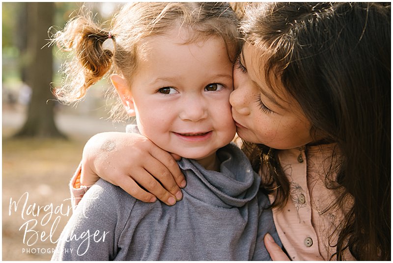 Close-up photo of big sister kissing her little sister on the cheek during their fall family session at Endicott Park in Danvers.