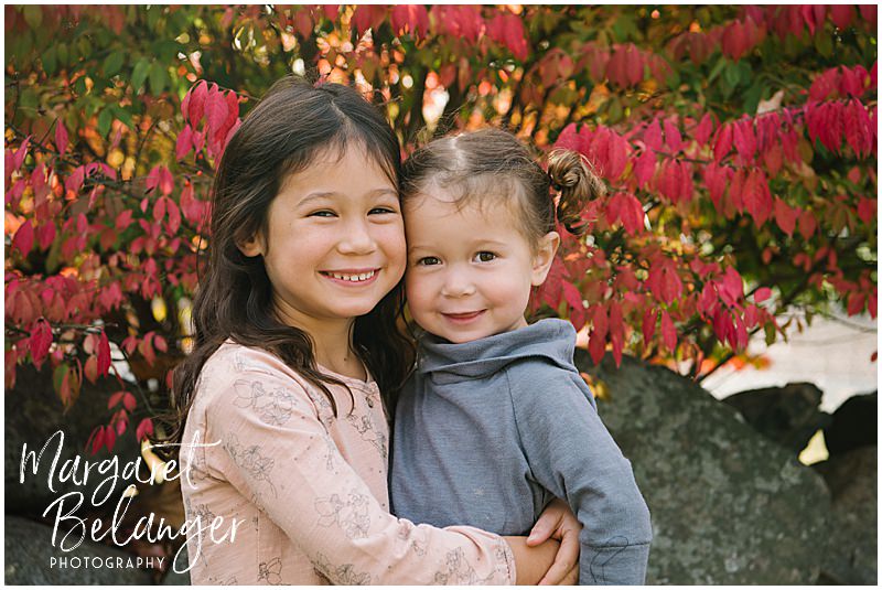 Two sisters hug in front of red leaves during their fall family session at Endicott Park in Danvers.
