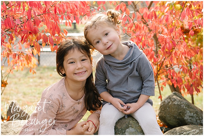 Two sisters sit on a rock wall and tilt their heads towards each other with red leaves framing them during their fall family session at Endicott Park in Danvers.