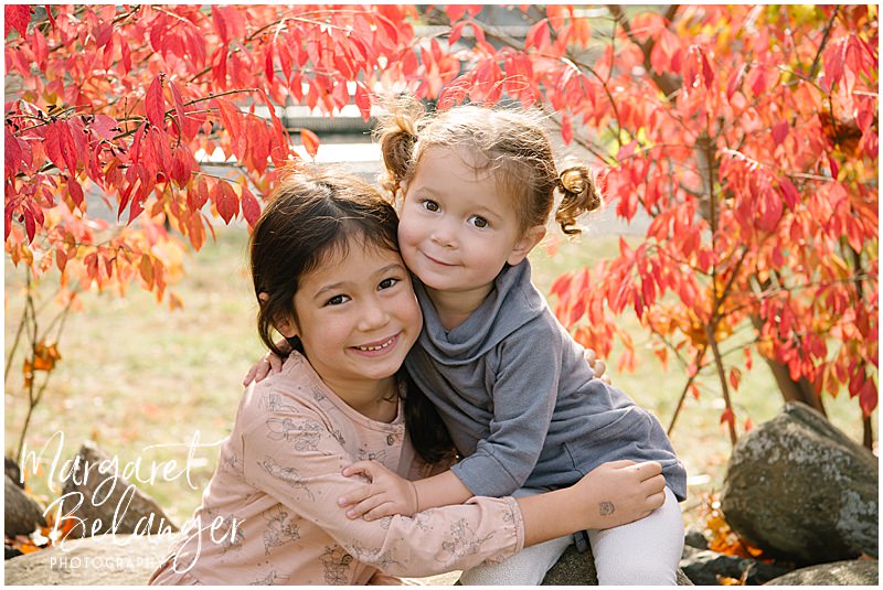 Two sisters sit on a rock wall and hug with red leaves framing them during their fall family session at Endicott Park in Danvers.