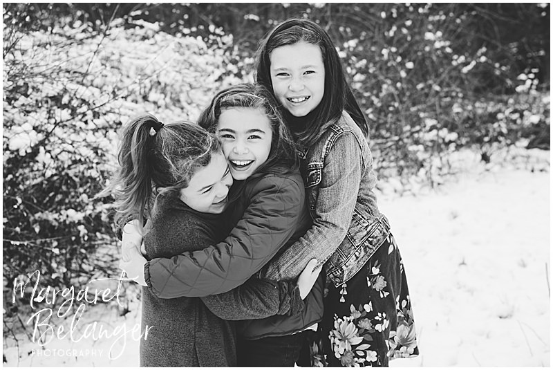 Black and white photo of three sisters huddling together and smiling for the camera at their winter family session at Minute Man National Park in Lincoln.