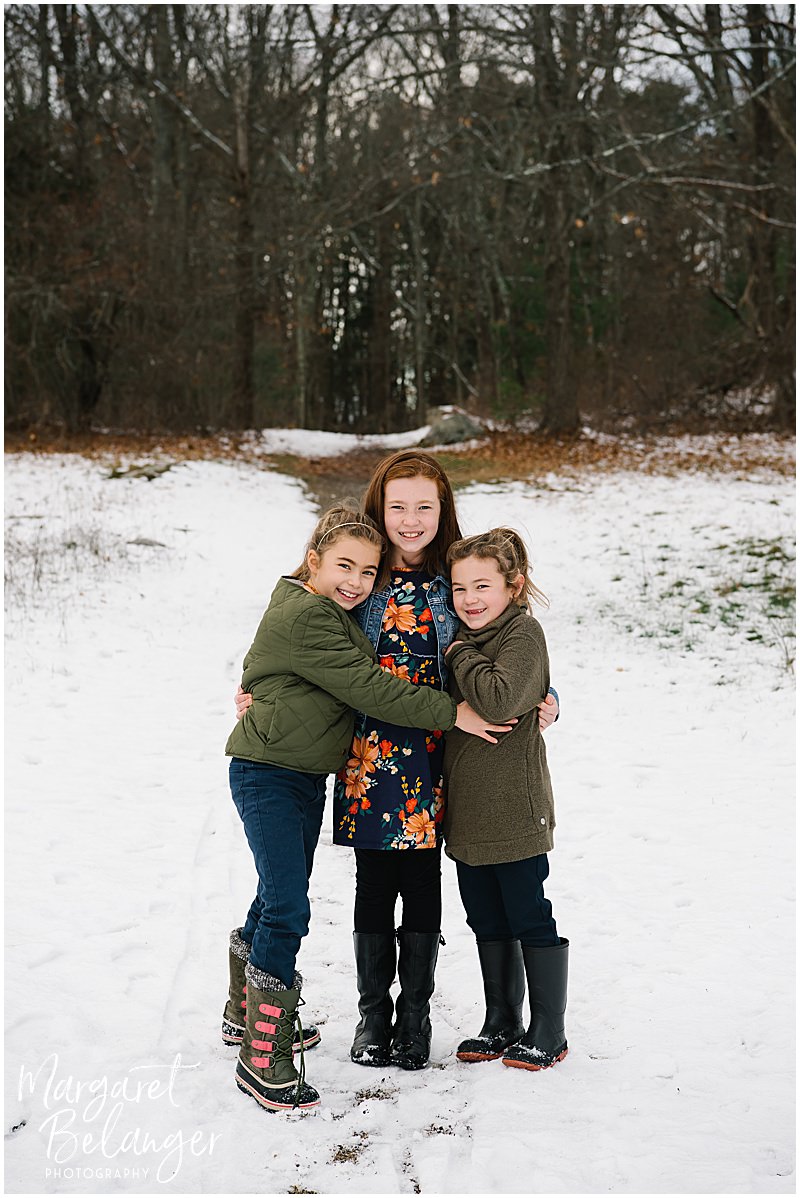 Three sisters standing together and hugging in the snow during their winter family session at Minute Man National Park in Lincoln.