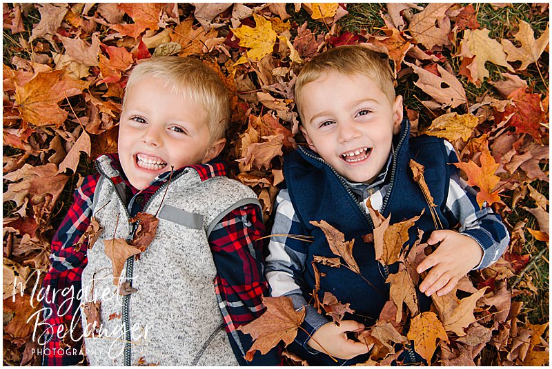 Two little boys smiling while lying in the leaves during their fall family session at Minute Man National Park in Lincoln, MA.