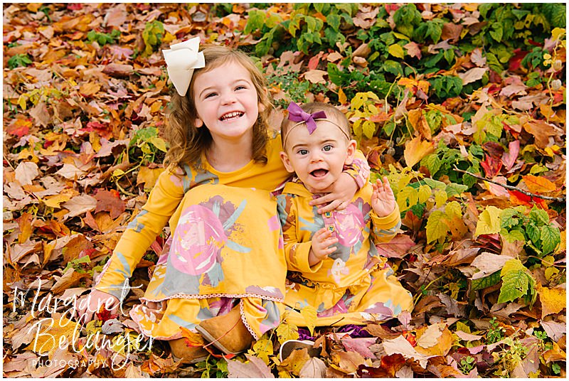 Two little girls hug and smile in the leaves at Mistletoe Christmas Tree Farm in Stow.