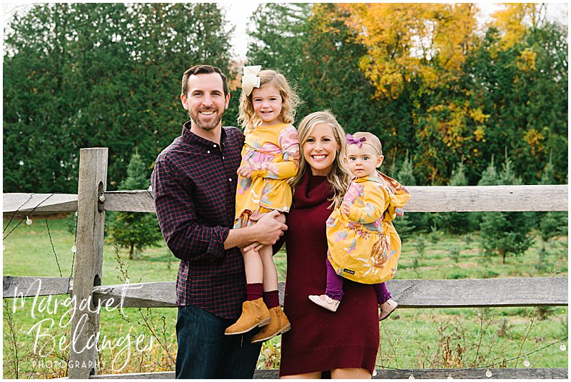 Mom and Dad hold their two daughters while standing in front of a wooden fence during their fall family session at Mistletoe Christmas Tree Farm in Stow.