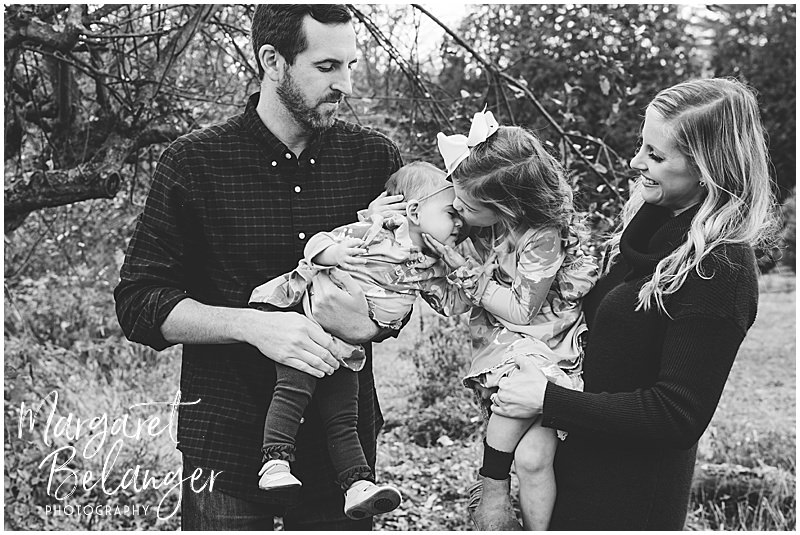 Mom and Dad hold their daughters while the big sister leans over and kisses her baby sister during their fall family session at Mistletoe Christmas Tree Farm in Stow.