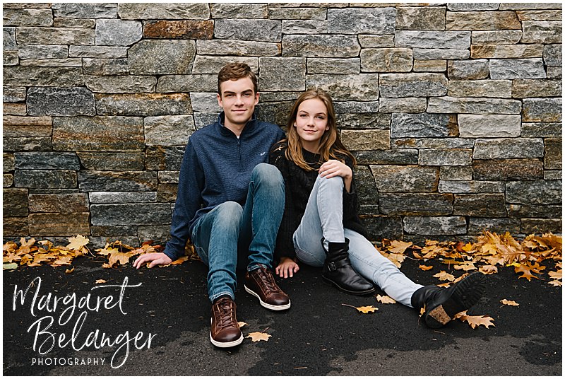 Teenage siblings sit on the driveway and lean against a stone wall during their Newton backyard family session.