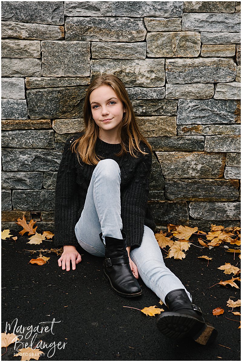 A teenage girl sits on the driveway and leans against a stone wall during a Newton backyard family session.