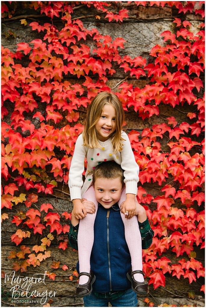 Little girl sitting on her big brother's shoulders in front of a wall of red leaves during their Winchester Center fall family session.
