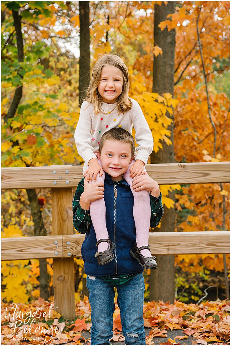 Little girl sitting on her brother's shoulders in front of a wooden fence during their Winchester Center fall family session.