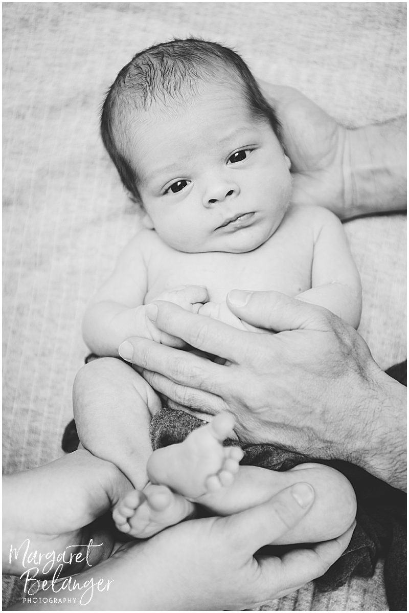 Black and white photo of a newborn boy looking at the camera with mom and dad's hands holding him.