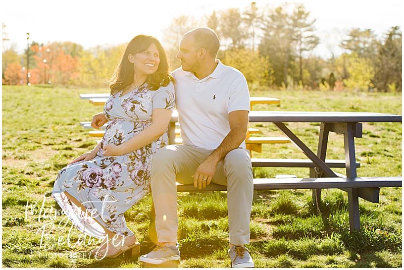 A couple looks at each other while sitting at a picnic table during their Nofolk, MA maternity session.