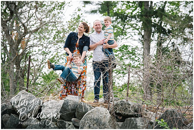 Family of 4 stands on a rocky hill, one son climbing a fence, for a family photo during their Minute Man National Park family session in Concord.