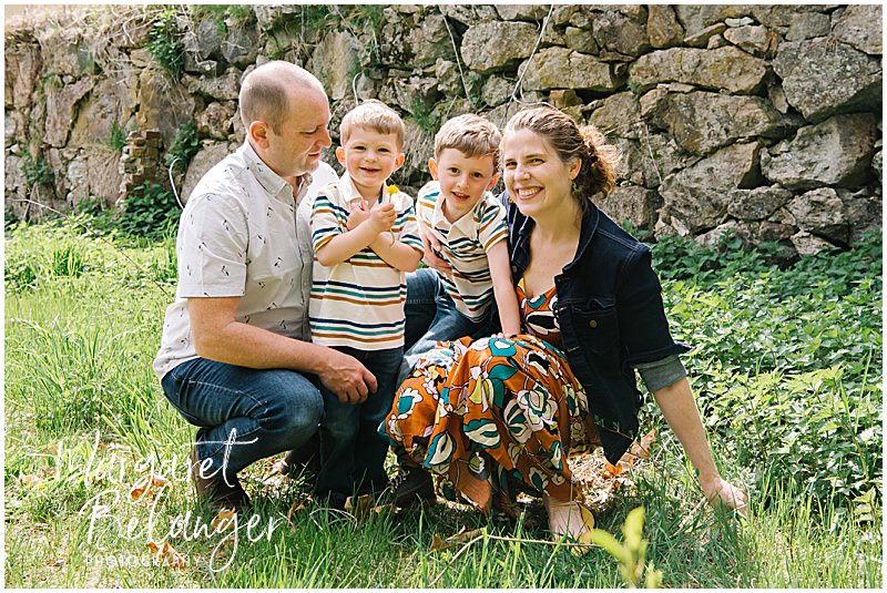 A family crouches down in the grass in front of a stone wall and poses together for a family portrait during a Concord family session at Minute Man National Park.