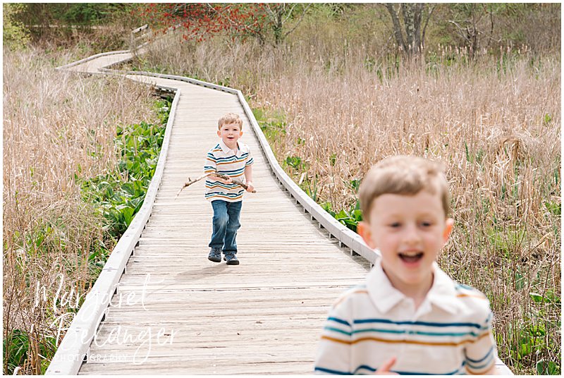 Two little boys, the youngest holding a big stick, run on a boardwalk at the Brooks Village area of Minute Man National Park during their spring family photo session in Concord.