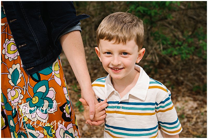 A little boy smiles for the camera while he holds his mom's hand during his Minute Man National Park family photo session in Concord.