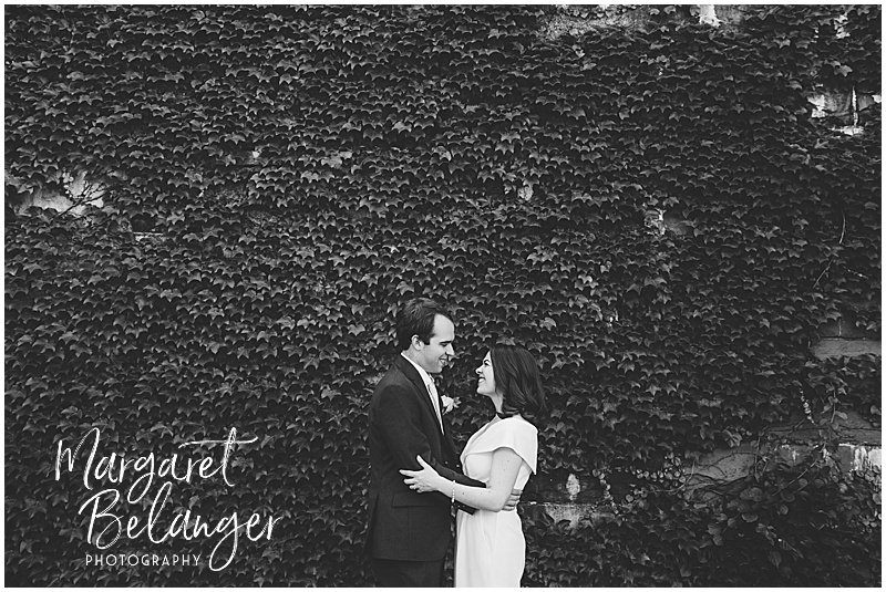 Black and white portrait of a bride and groom standing in front of a green vine-covered stone wall in Winchester Center.
