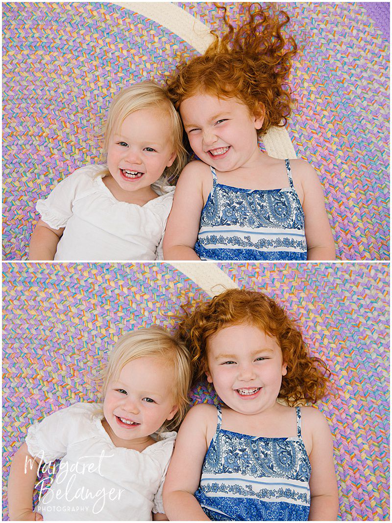 Two little girls, a blonde and a redhead, lie on a colorful braided rug with their heads close together.