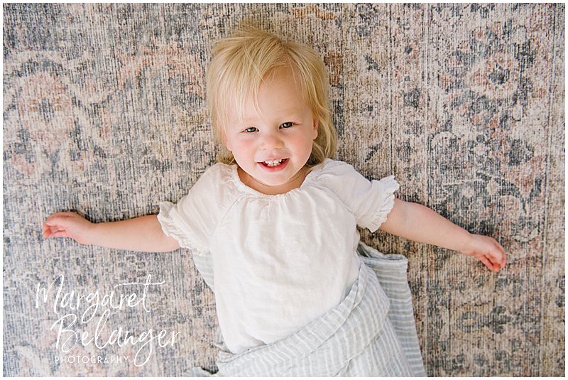 A little blonde girl lies on a colorful rug and smiles for the camera.