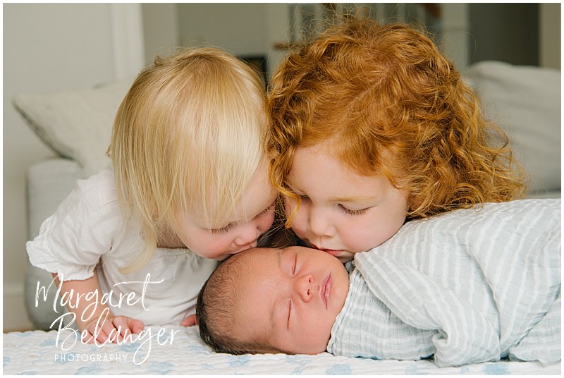 A little blonde girl and a little redheaded girl lean in and kiss their brunette newborn baby brother during a Winchester, MA newborn session.