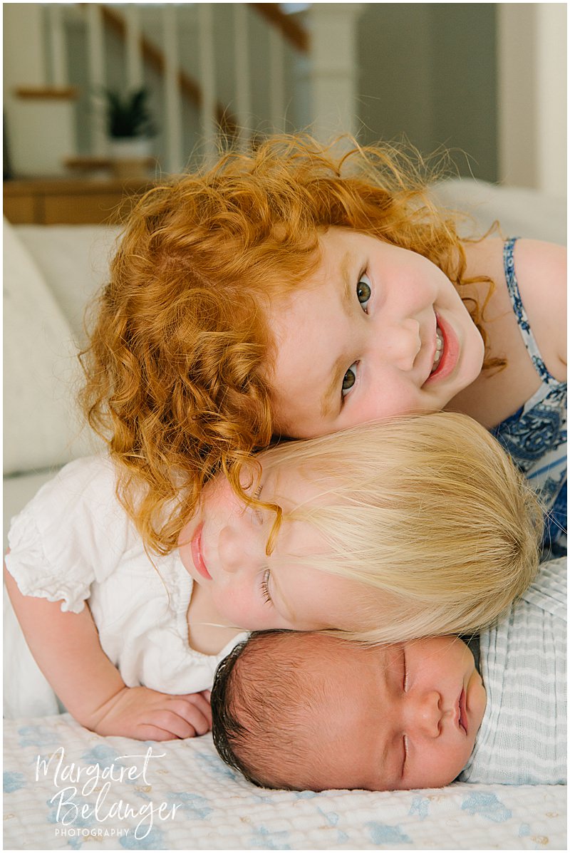 A brunette newborn boy sleeps on a blanket while his blonde sister puts her head on his and then a redheaded sister puts her head on top.