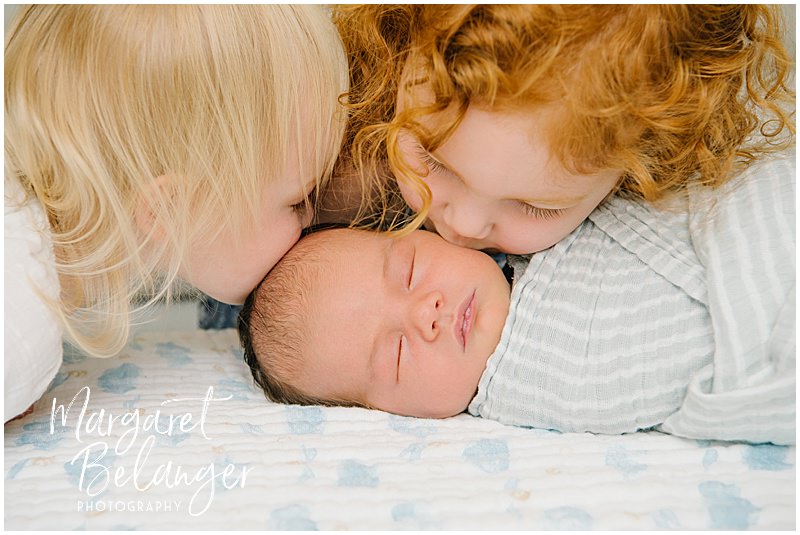 A little blonde girl and a little redheaded girl lean in and kiss their brunette newborn baby brother during a Winchester, MA newborn session.