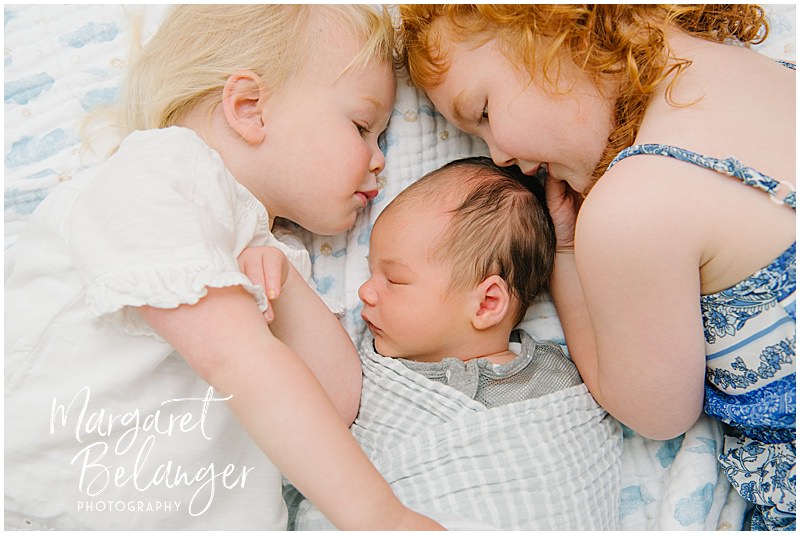 A newborn baby boy lies on a blanket while his sisters lie next to him, leaning their heads in towards his.