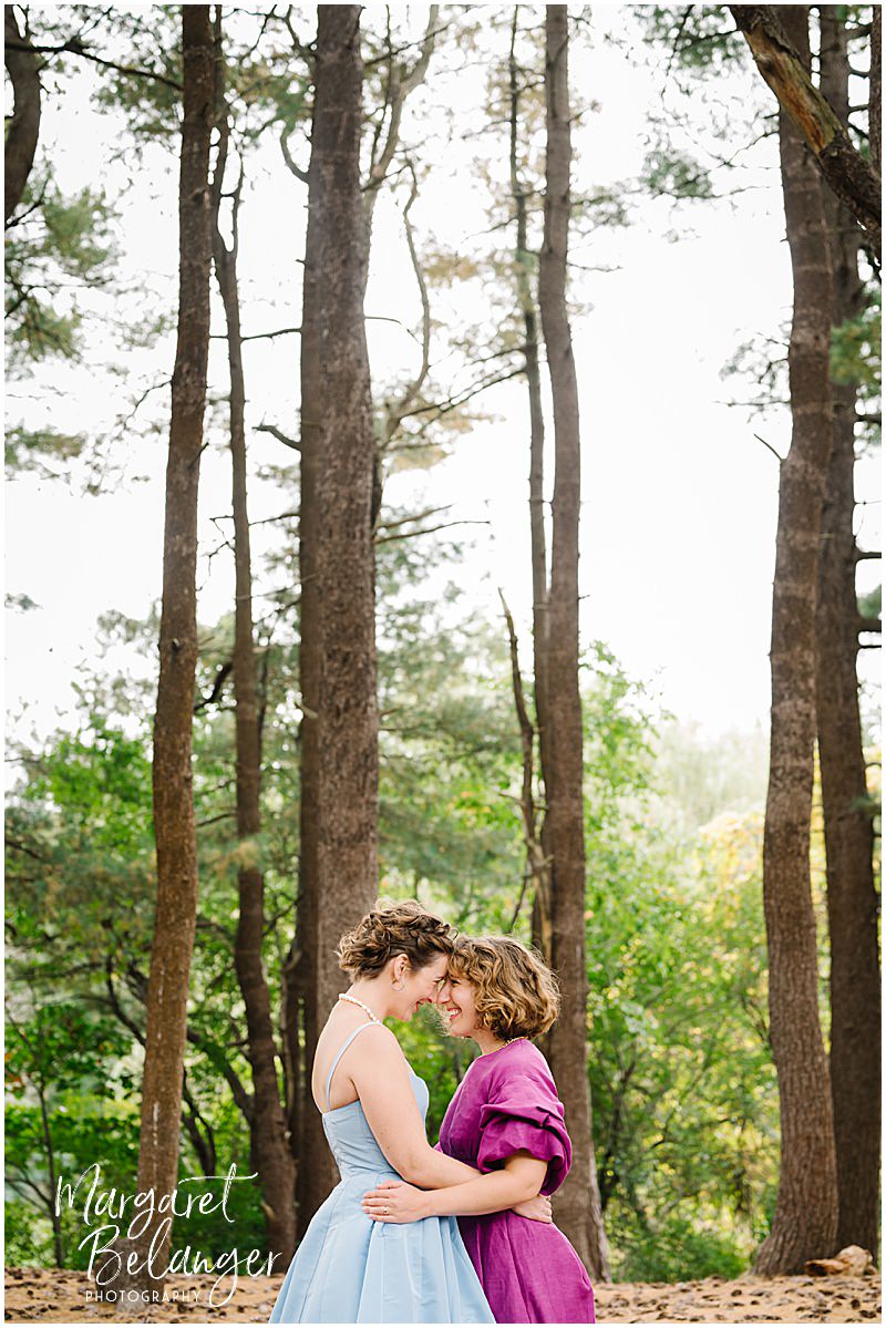 Two brides lean into each other and smile among the trees at Allandale Farm in Brookline