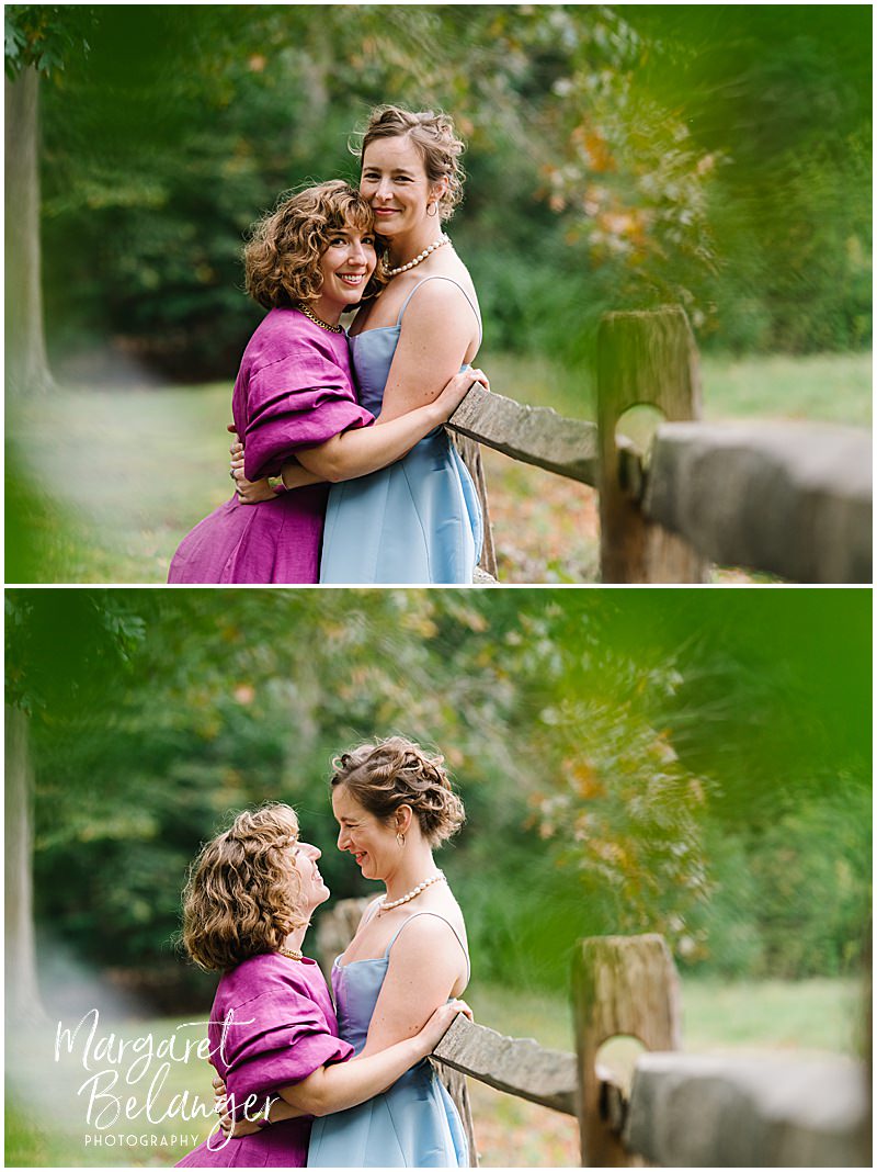 Two brides in non-traditional wedding dresses hugging in front of a fence.