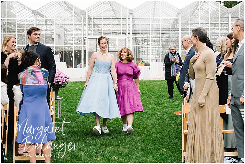 Two brides walk down the aisle with a greenhouse behind them.