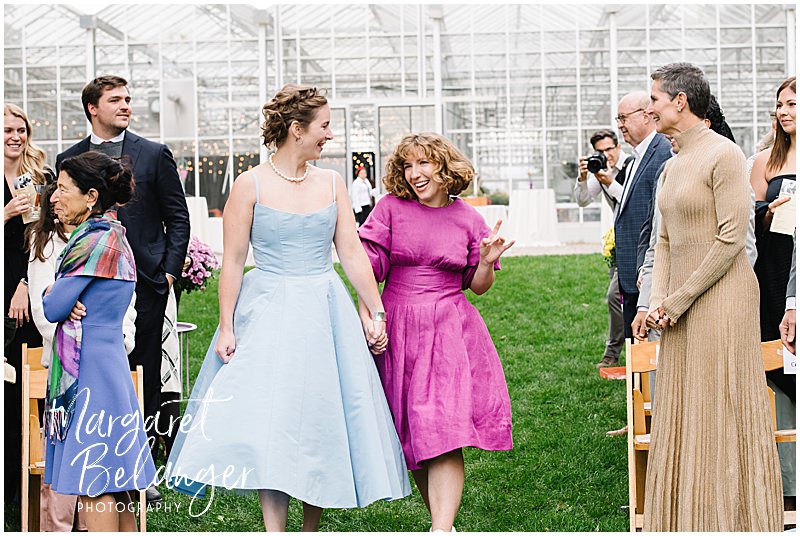 Two brides dance down the aisle in front of a greenhouse.