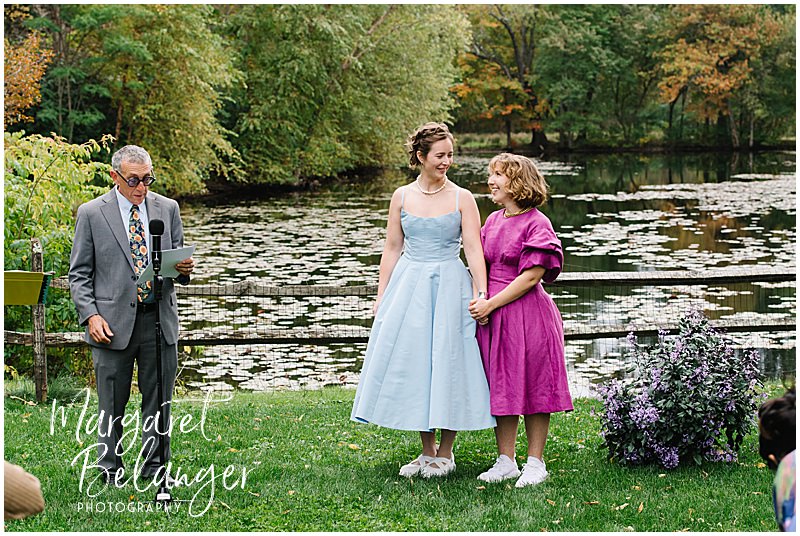 Two brides stand in front of a pond during their wedding ceremony.
