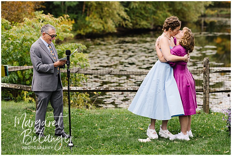 Brides share their first kiss in front of a pond during their wedding ceremony.