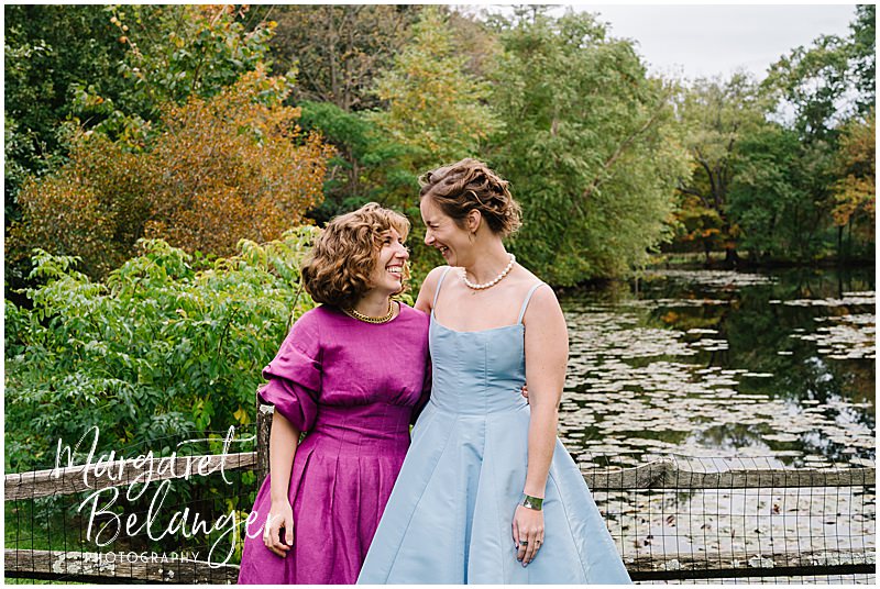 Brides smile at each other while they lean against a fence in front of a pond.