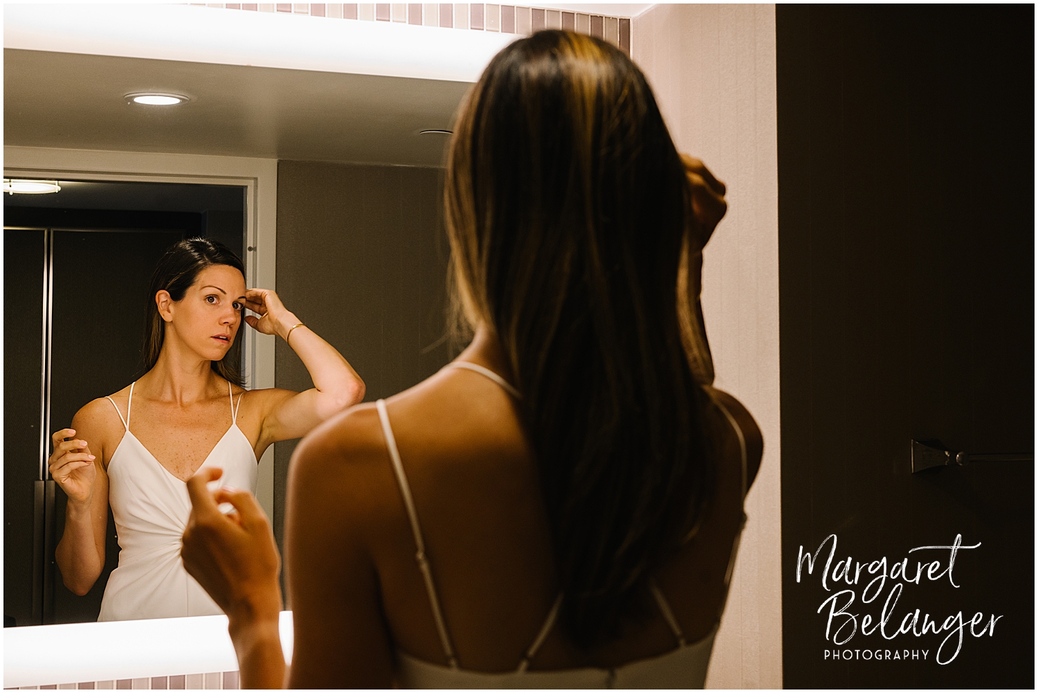A bride in a white dress looks at her reflection in a mirror, adjusting her hair.