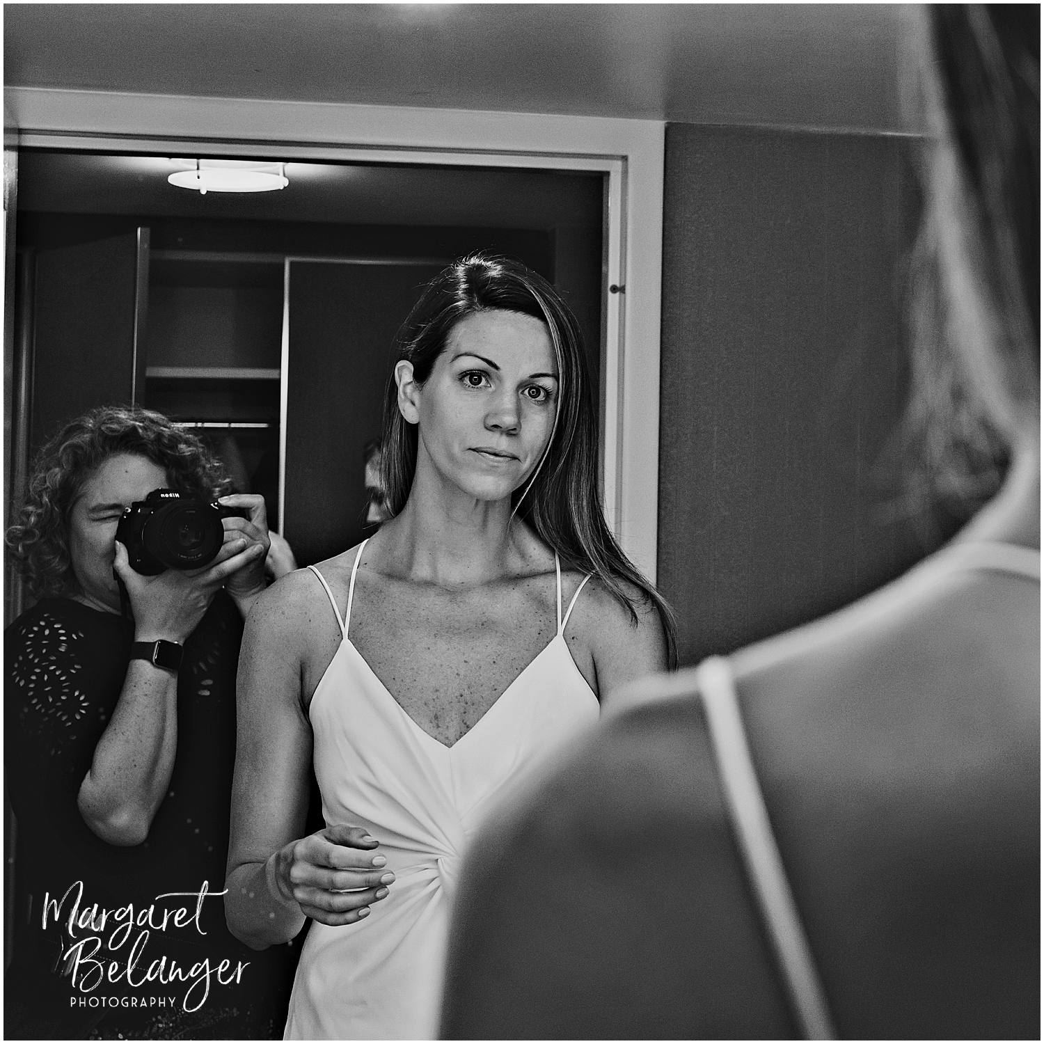 A bride in a white dress looks into a mirror while a photographer captures the moment.