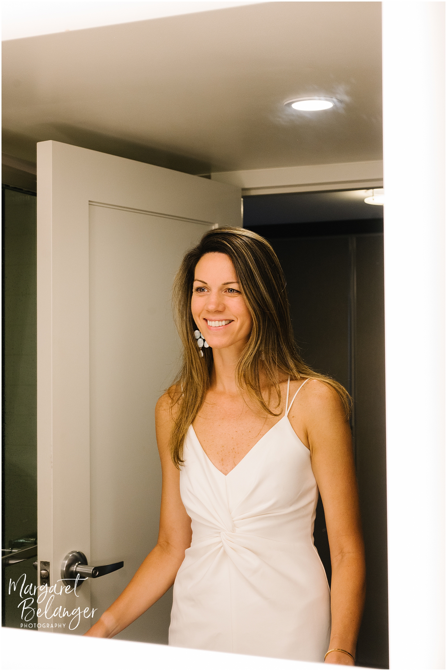 A smiling bride in a white dress standing in front of a mirror.