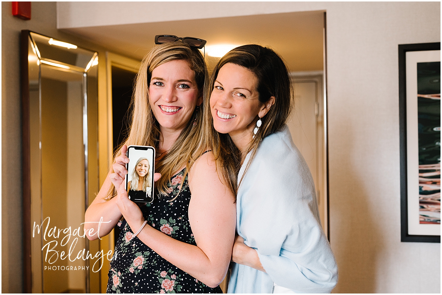 The bride and her sister posing for a photo, one holding a smartphone with their other sister on FaceTime.