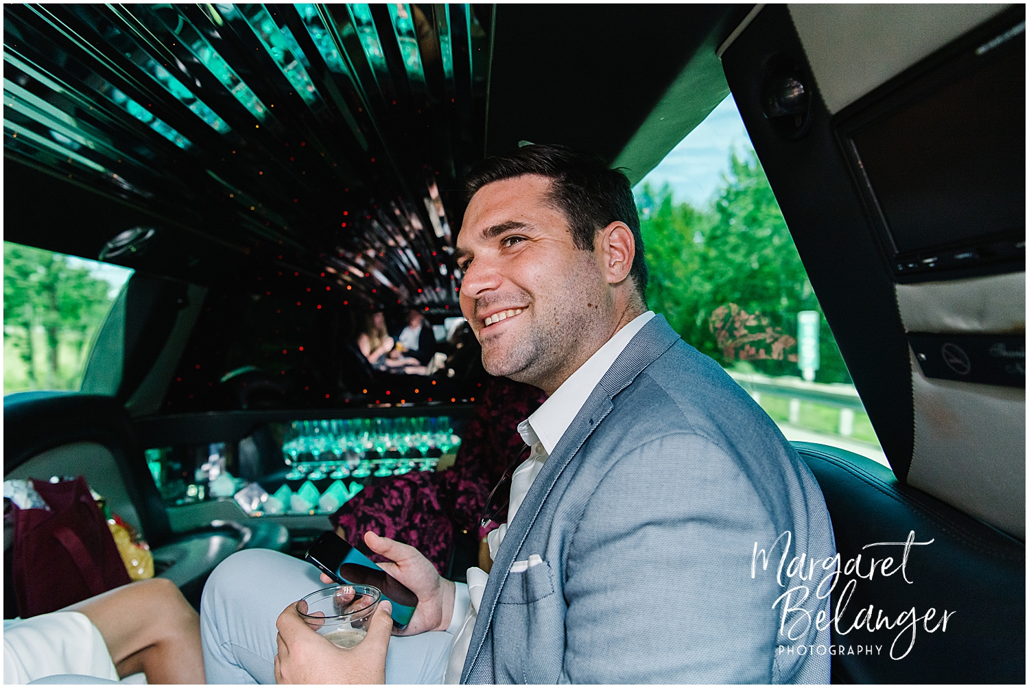 A man in a grey suit smiling inside a limousine with colorful lights on the ceiling.