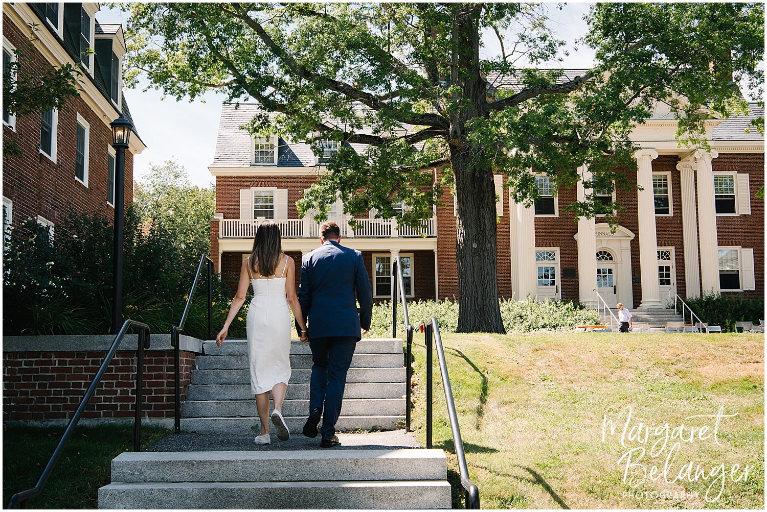 A bride and groom holding hands while walking up the steps towards a traditional brick building on a sunny day.