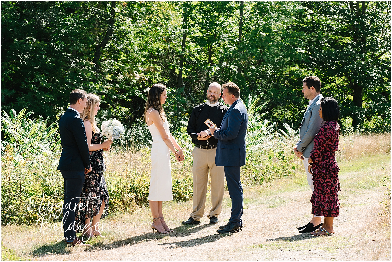 Outdoor wedding ceremony with a couple exchanging vows, officiant and attendees watching.