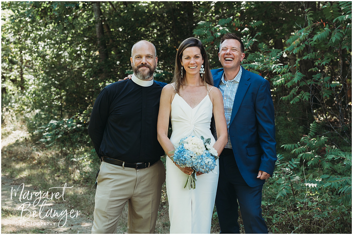 A bride in a white dress holding a bouquet stands between two smiling men, one in a clerical collar and the other in a blue suit, outdoors.